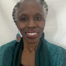 A smiling therapist from Total Life with short, braided gray hair, wearing vibrant earrings and a green shawl, photographed against a neutral backdrop."