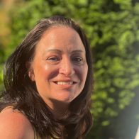 "A smiling therapist from Total Life with shoulder-length dark brown hair, wearing a yellow sleeveless top, photographed outdoors with lush greenery in the background.