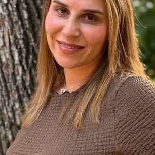 A smiling therapist from Total Life with straight blonde hair, wearing a brown textured top, photographed outdoors beside a tree with greenery in the background.