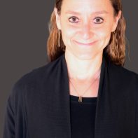 A therapist from Total Life with long, light brown wavy hair, wearing a black outfit and gold earrings. She is smiling warmly and has a professional appearance, complemented by a neutral background that enhances her approachable demeanor.