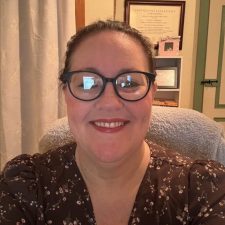 "A smiling therapist from Total Life with glasses and pulled-back hair, wearing a floral-patterned blouse, photographed indoors with framed certificates and a cozy setting in the background.