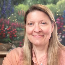 A therapist from Total Life with a friendly expression, dressed in a light pink top. She is positioned in front of a vibrant background showcasing a garden scene with colorful flowers, creating a serene and inviting atmosphere.