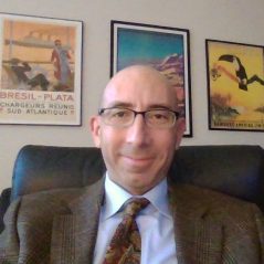 A therapist from Total Life seated in an office setting. He is wearing a suit with a patterned tie and glasses, with framed travel-themed posters adorning the wall behind him. The overall ambiance is professional and approachable.