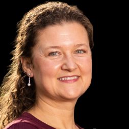 A professional headshot of a therapist from Total Life, a smiling woman with curly hair, wearing pearl earrings and a burgundy top, set against a solid black background.