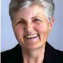 A smiling therapist from Total Life with short gray hair, wearing a black blazer and a light-patterned blouse, photographed against a plain light blue background.