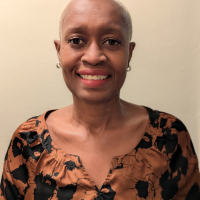 A professionally dressed therapist at Total Life with short silver hair and a smile, wearing a patterned orange and black blouse, standing against a neutral beige background.