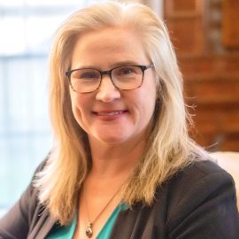 A smiling therapist from Total Life with blonde hair, wearing glasses, a teal top, and a black blazer, accessorized with a pendant necklace, photographed indoors with a softly lit background featuring wooden decor and a window.