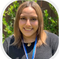 Bright outdoor portrait of a smiling therapist at Total Life with shoulder-length light brown hair, wearing a black top and a blue lanyard, accessorized with layered gold necklaces, set against a backdrop of green foliage.