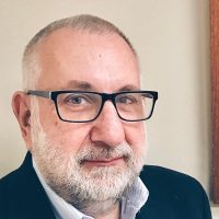 A therapist from Total Life with short gray hair, a beard, and wearing black glasses, a light blue shirt, and a dark blazer, standing in front of a neutral background.