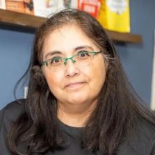 A Total Life therapist with long dark hair, wearing glasses with green frames and a dark top, standing in front of a blue wall with shelves holding books and framed artwork.