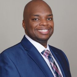 A therapist from Total Life dressed in a professional blue suit with a white shirt and a plaid tie. He has a bright smile, a shaved head, and a light gray background, conveying a polished and approachable demeanor.