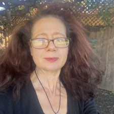 A therapist from Total Life with voluminous auburn hair, wearing glasses, a black outfit, and a lotus-shaped pendant necklace, photographed outdoors with trees and a wooden fence in the background.