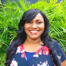 A therapist from Total Life with long, wavy black hair, wearing a floral patterned top. She is smiling brightly against a vibrant green background of lush plants, which adds a fresh and welcoming feel to the image.