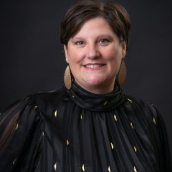 A smiling therapist from Total Life with short brown hair, wearing a black top with gold accents and large gold earrings, photographed against a dark background.