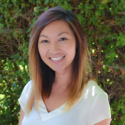 A therapist from Total Life with medium-length, ombre-style hair, smiling warmly in an outdoor setting with greenery in the background. She is wearing a white top, exuding a professional and approachable demeanor.