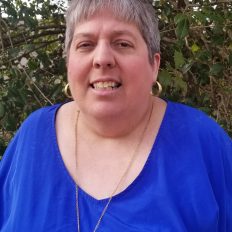 A smiling therapist from Total Life with short gray hair, wearing gold earrings, a necklace, and a bright blue top, photographed outdoors with greenery in the background.