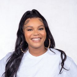 A smiling therapist from Total Life with long wavy black hair styled in a high ponytail, wearing large gold hoop earrings and a white top, photographed against a plain white background.