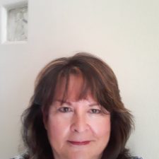 A therapist from Total Life with shoulder-length brown hair, smiling softly against a light-colored wall backdrop. She is wearing a patterned top, projecting a professional and warm presence.