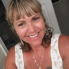 A therapist from Total Life with a radiant smile and welcoming demeanor. She is dressed in a floral-patterned top and accessorized with layered gold necklaces and earrings, creating a warm and approachable appearance. The background includes elements of a cozy, well-lit room, enhancing the personal and inviting tone of the portrait.