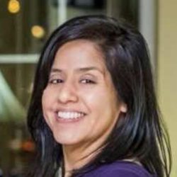 A therapist from Total Life with shoulder-length black hair, smiling brightly in an indoor setting. She is wearing a purple top and appears cheerful and approachable, reflecting a professional demeanor.