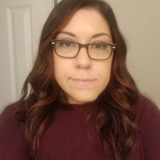 A therapist from Total Life with shoulder-length dark hair with red highlights, wearing glasses and a maroon sweater, standing indoors in front of a neutral-colored wall and door.