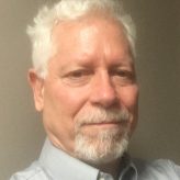 A therapist from Total Life, exuding confidence and professionalism. He has neatly styled silver hair and a trimmed beard, wearing a light-colored collared shirt that complements his approachable demeanor. The neutral background keeps the focus on the subject, highlighting his thoughtful and composed expression.