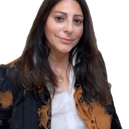 A therapist from Total Life. She is dressed in a stylish, tie-dyed jacket over a white shirt, accessorized with a gold pendant necklace. Her dark hair is worn down, and she smiles gently at the camera against a white background.