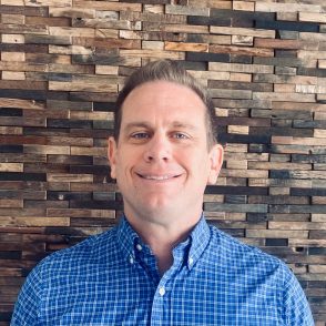 A professional headshot of a man with short hair, a therapist at Total Life, wearing a blue checkered button-up shirt, smiling warmly. The background is composed of a textured wooden wall, adding a natural and modern feel.