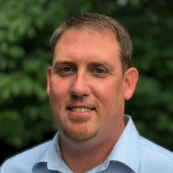 A therapist from Total Life with short brown hair and a light beard, wearing a light blue collared shirt, smiling in front of a blurred background of green foliage.