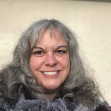 A therapist at Total Life with wavy gray hair and a warm, cheerful smile. She is wearing a patterned scarf and is photographed outdoors against a neutral beige background, enjoying natural sunlight.