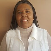 A smiling therapist from Total Life with braided hair, wearing a white sweater over a turtleneck, photographed indoors against a beige background.