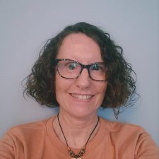 A smiling therapist from Total Life with curly brown hair, wearing glasses, a necklace with a pendant, and an orange shirt, photographed against a light blue background.