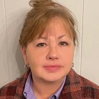 A therapist from Total Life with light brown hair tied in a bun, wearing a purple blouse and a plaid jacket, posing against a pale-colored wall.