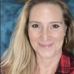 Close-up portrait of a smiling therapist at Total Life with long, light blonde hair, wearing a red plaid shirt, and gold hoop earrings, set against a soft, blurred indoor background with shades of blue.