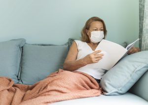 older woman with medical mask home during pandemic reading book