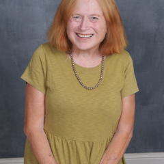 A smiling therapist from Total Life with shoulder-length red hair, wearing a green dress and a pearl necklace, photographed indoors against a gray textured background.