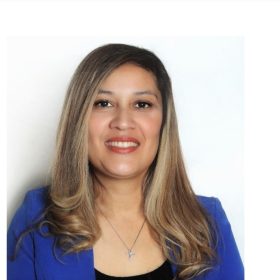 A therapist at Total Life with long blonde hair styled in soft waves, wearing a royal blue blazer and a silver cross necklace. She is smiling warmly against a clean white background.