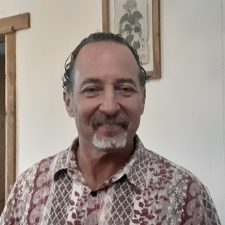 A therapist at Total Life with short dark hair and a mustache, wearing a patterned shirt. He is smiling warmly in an indoor setting with light-colored walls and wooden door frames in the background.