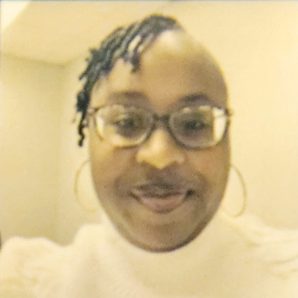 A therapist at Total Life with short curly hair and glasses, wearing a white sweater. She is smiling warmly in a neutral indoor setting, with soft lighting in the background.