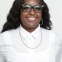 A therapist at Total Life with long dark hair styled in soft waves, wearing a white button-down shirt and a gold necklace. She is smiling warmly against a simple white background.