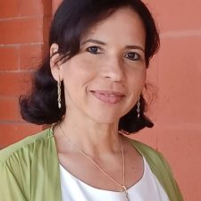 A therapist at Total Life with dark hair styled in loose waves, wearing a white top with a light green cardigan and a necklace. She is smiling warmly in an outdoor setting with a brick wall in the background.