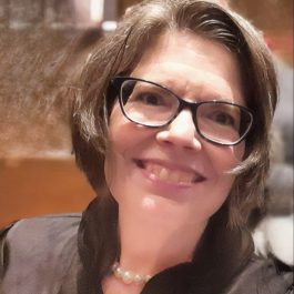 A therapist at Total Life with short brown hair and glasses, wearing a black top and a pearl necklace. She is smiling warmly in an indoor setting with a blurred background.