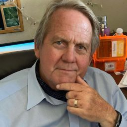 A therapist at Total Life with short blonde hair, wearing a light blue dress shirt and a dark patterned neck scarf. He is seated in an office setting, resting his chin on his hand with a thoughtful expression. Behind him, a desk with office supplies and a bulletin board with photographs are visible.