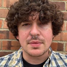 A therapist at Total Life with curly brown hair, wearing a plaid shirt over a black t-shirt and a septum piercing. They are standing against a brick wall in an outdoor setting with a neutral expression.