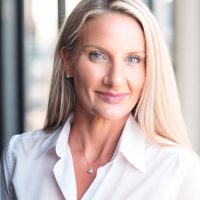 A Total Life therapist with long blonde hair, wearing a white shirt, smiling gently at the camera with a blurred city background.