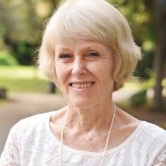 A senior woman with short blonde hair, wearing a white lace top, smiling warmly at the camera outdoors with a green, peaceful background.